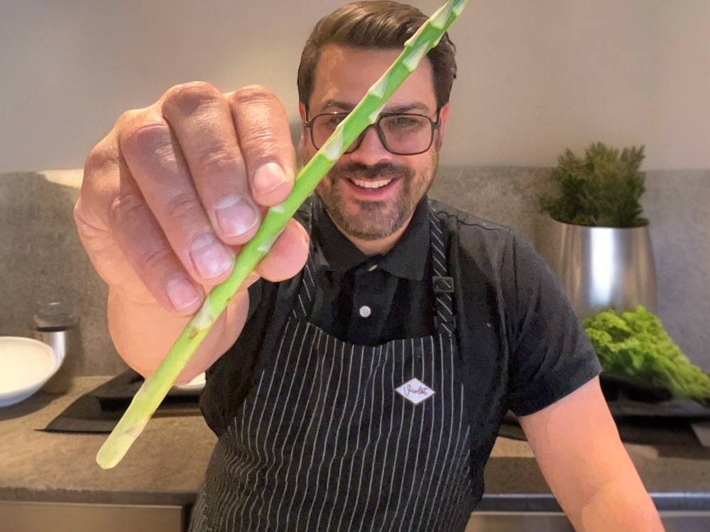 Jesse Brune grinning in a striped apron, holding a spear of asparagus up to the camera.