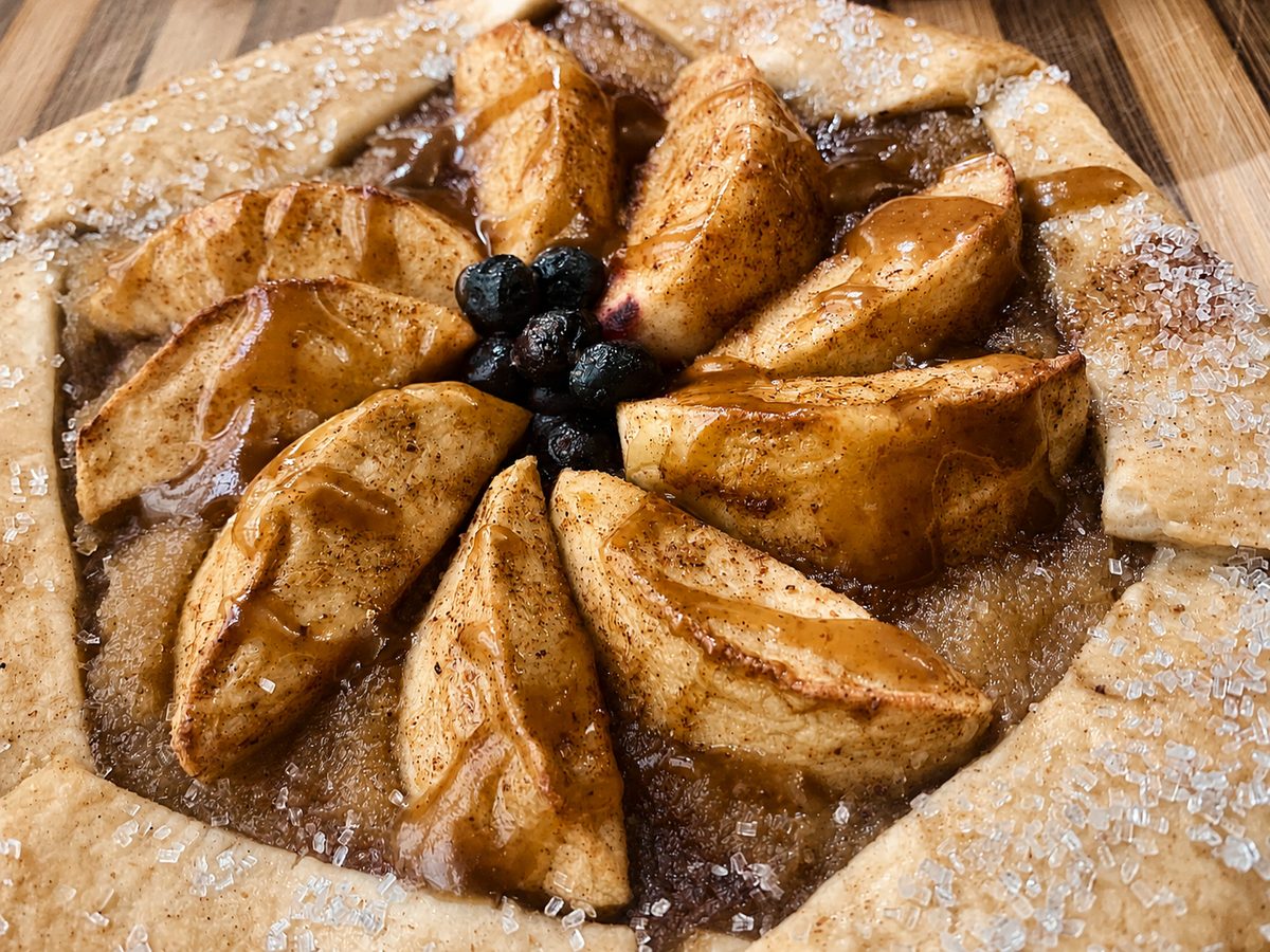 A rustic apple galette with blueberries at the centre, cinnamon, a caramel drizzle and a sugared crust.