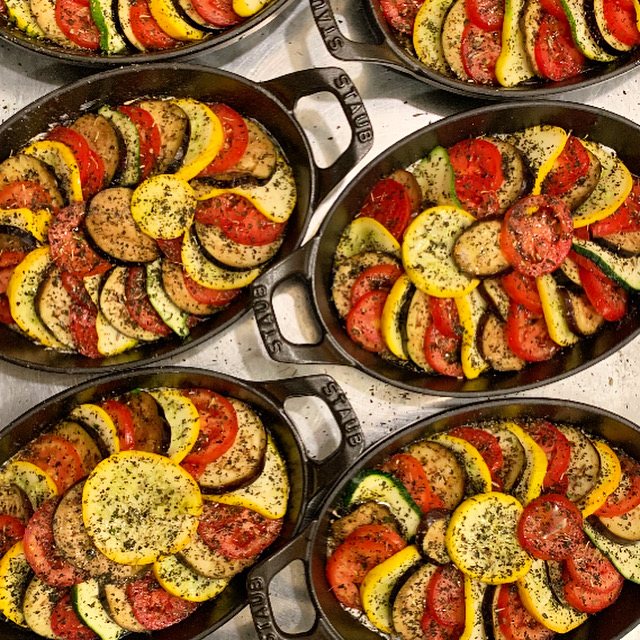 Six oval cast iron dishes of ratatouille, sliced tomato, zucchini, squash and eggplant shingled in tight spirals with herbs.